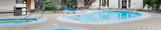 Indoor pool area with multiple circular pools. Lounge chairs are placed around the pools, and towels are neatly stacked on a rack. Large windows and ceiling beams enhance natural light. A plant and stairs leading to a mezzanine level are visible.