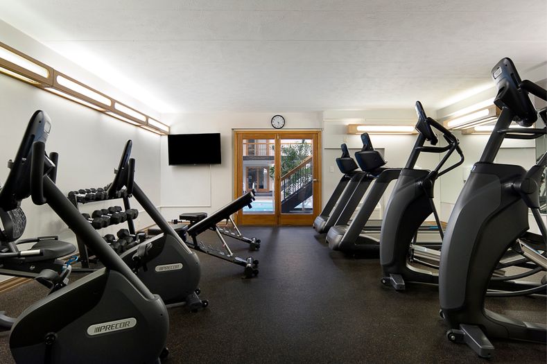 A bright, modern gym with exercise bikes, a treadmill, and a bench facing a flat screen TV. A clock hangs above a wooden door leading outside, revealing a glimpse of trees and a building.