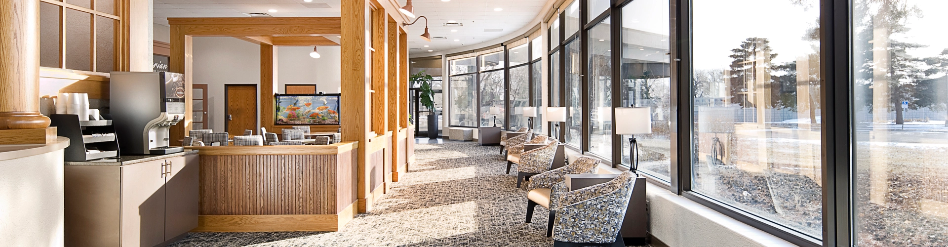 Bright, spacious room with large windows, patterned carpet, and wooden accents. Features a kitchenette area with coffee machines on the left and a seating area with patterned chairs and lamps on the right, overlooking a sunlit outdoor scene.