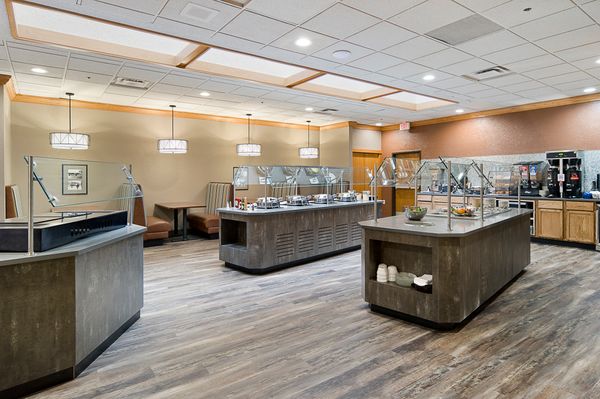 A spacious cafeteria with several food stations featuring an assortment of dishes. There are wooden floors, modern light fixtures, and booth seating. Various kitchen appliances are visible in the background.