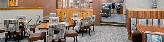 Restaurant interior with patterned chairs around square tables on a carpeted floor. Booth seating is on the right, and a decorative plant stands beside a hanging light fixture. In the background, there's a buffet area and wooden paneling.