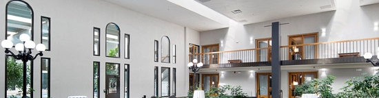 Spacious indoor event area with high ceilings, arched windows, and multiple round tables with white tablecloths and black chairs. A balcony overlooks the space, and potted plants are placed around the room.