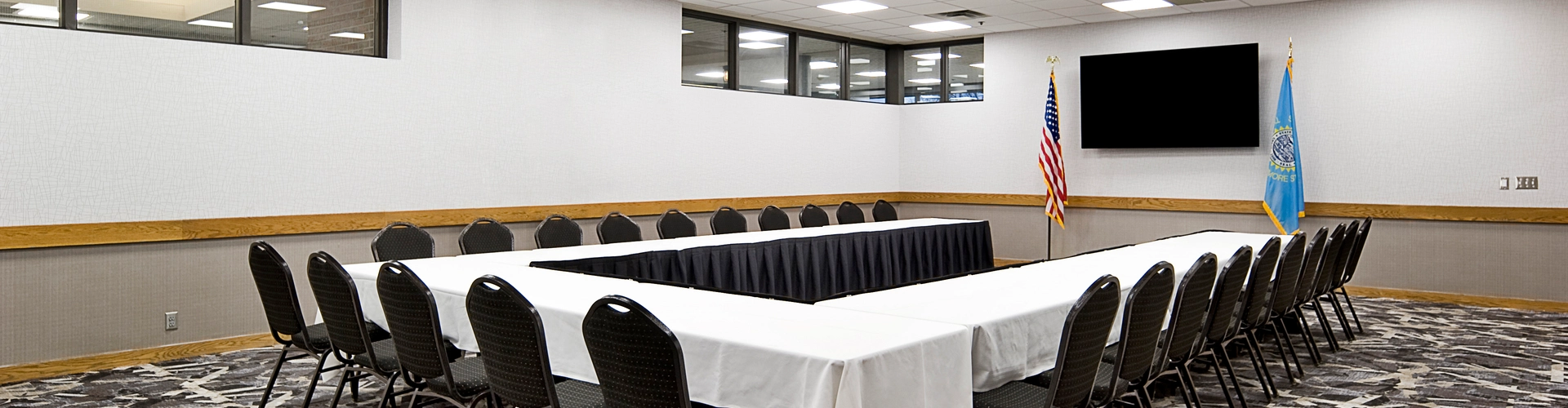 A conference room with an empty U-shaped table setup. The table is covered with white tablecloths and surrounded by black chairs. Flags and a large flat-screen TV are on the wall. The carpet has a geometric pattern, and the ceiling has recessed lighting.