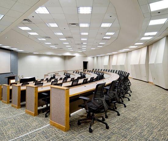 A curved lecture hall with rows of empty swivel chairs and desks facing a podium and dual projection screens. The carpeted floor and ceiling lights create a well-lit, modern classroom environment.
