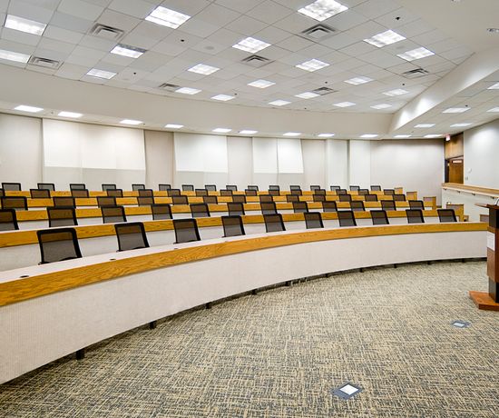 An empty lecture hall with rows of tiered seating featuring wooden desks and black mesh chairs. A podium with a microphone stands at the front. The ceiling has a grid of recessed lights, and the walls are plain.