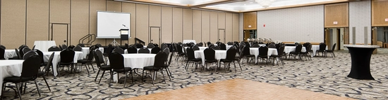 A spacious, empty banquet hall with round tables covered in white tablecloths and surrounded by black chairs. There's a podium and projection screen in the background, with large chandeliers and a patterned carpet on the floor.