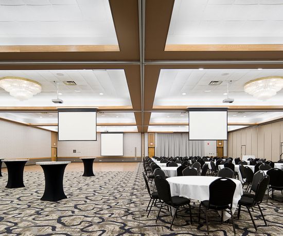 A spacious banquet hall with round tables covered in white tablecloths and black chairs arranged on a patterned carpet. Two projectors are set up, and elegant chandeliers hang from the ceiling. Black-covered tall cocktail tables are also present.