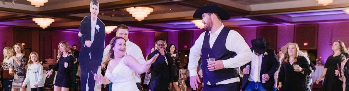 A bride in a white gown dances with people dressed in Western attire on a wooden dance floor. Guests in the background are dancing near a life-sized cutout of a person in formal wear. The room is lit with warm, purple-tinted lights.