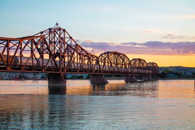 A long, iron railway bridge stretches across a calm river under a colorful sunset sky. The structure is reflected in the water, with soft orange and pink hues in the sky complementing the scene.
