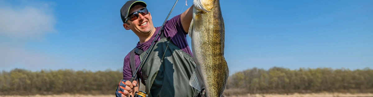 A person in fishing gear and sunglasses is standing in shallow water, holding up a large fish with a fishing rod. The background features a calm river and a tree line under a clear blue sky.