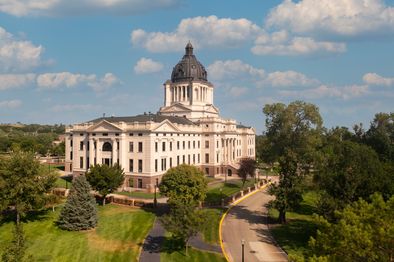 A large, historic government building with a central dome stands surrounded by trees and greenery. The building is white with intricate architectural details. A driveway curves in front, and the sky above is partly cloudy.