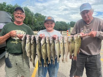 Three people smiling and holding a line of twelve fish in a parking lot. They appear happy and are standing under a cloudy sky. Trees are visible in the background.