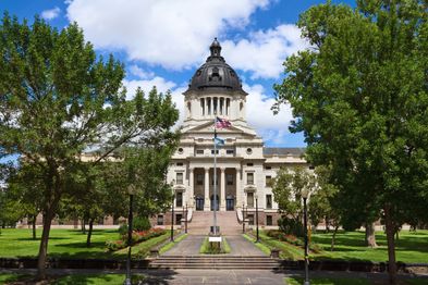 The image shows a large, historic building with a central dome and American flag, surrounded by trees and a well-maintained garden. A pathway leads to the entrance, flanked by lampposts. The sky is blue with scattered clouds.