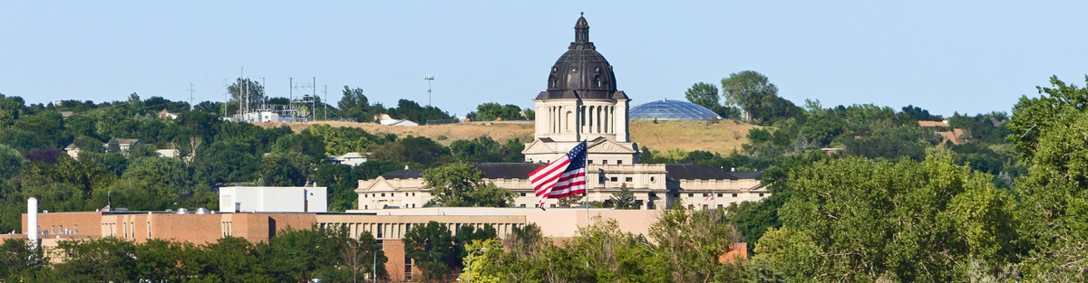 A distant view of the South Dakota State Capitol building with a large American flag displayed in front. The scene includes surrounding trees, a clear blue sky, and a body of water in the foreground.