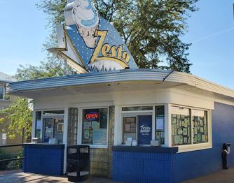 Small vintage ice cream stand with a retro "Zesto" sign featuring a cone. The building is blue and white with large windows displaying various colorful signs and posters. An "OPEN" sign is illuminated next to a tree and trash bin. Sky is clear.