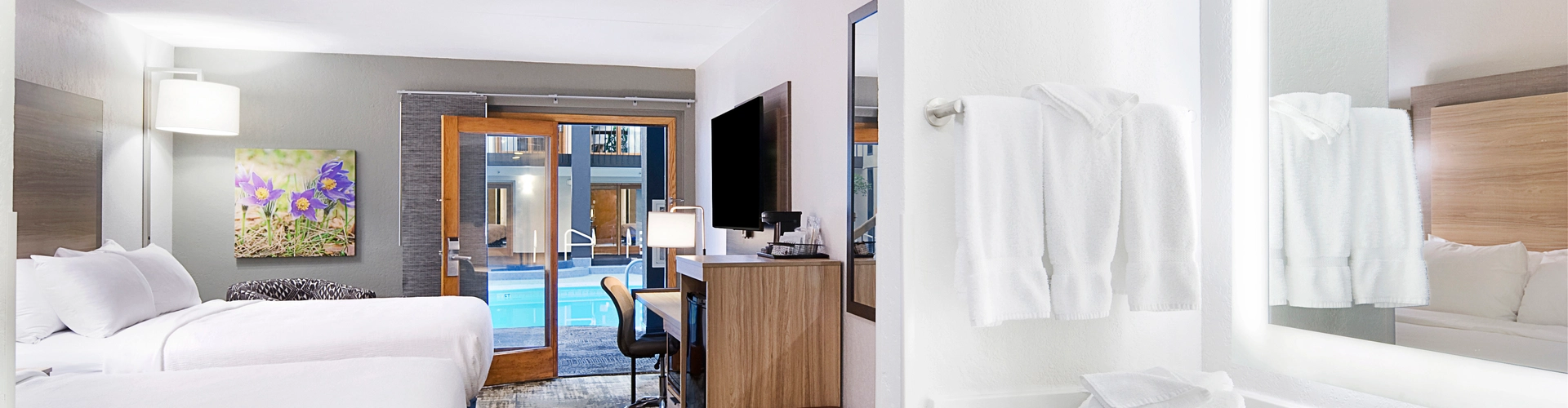 Modern hotel room with two beds featuring white linens, a work desk with a chair, a wall-mounted TV, and a bright bathroom area with a sink and mirror. A sliding glass door opens to reveal a pool view outside.