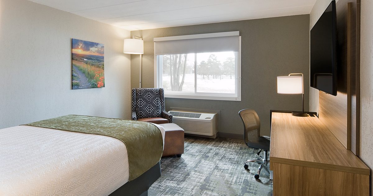 A modern hotel room with a large bed, green throw blanket, and a bedside lamp. There's a desk with a chair and lamp, a large TV mounted on the wall, a patterned armchair, and a window showing a snowy landscape outside.
