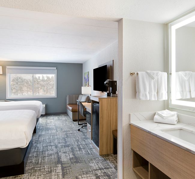 Modern hotel room with two beds and a view of trees through a window. There's a flat-screen TV, desk, and chair. A well-lit vanity with a sink and mirror is in the foreground, along with neatly folded towels. Carpeted floor and neutral decor.