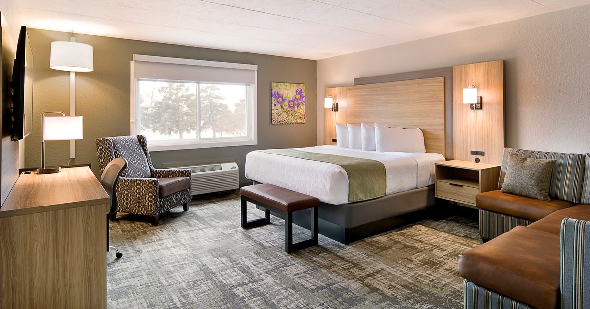 A modern hotel room featuring a king-size bed with white linens and a gray bed runner. There's a patterned armchair, a small sofa, and a bench. A large window with a view, a wall-mounted TV, and minimalistic decor complete the space.