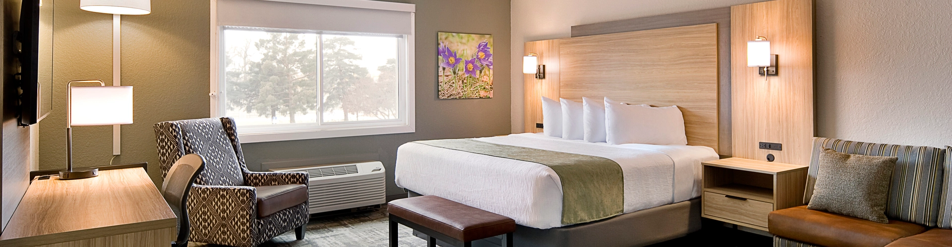 Modern hotel room with a king-size bed, wooden headboard, and bedside tables. An armchair is near the window, and artwork hangs on the wall. A bench sits at the foot of the bed, and a wall-mounted TV is across from it. Neutral tones throughout.