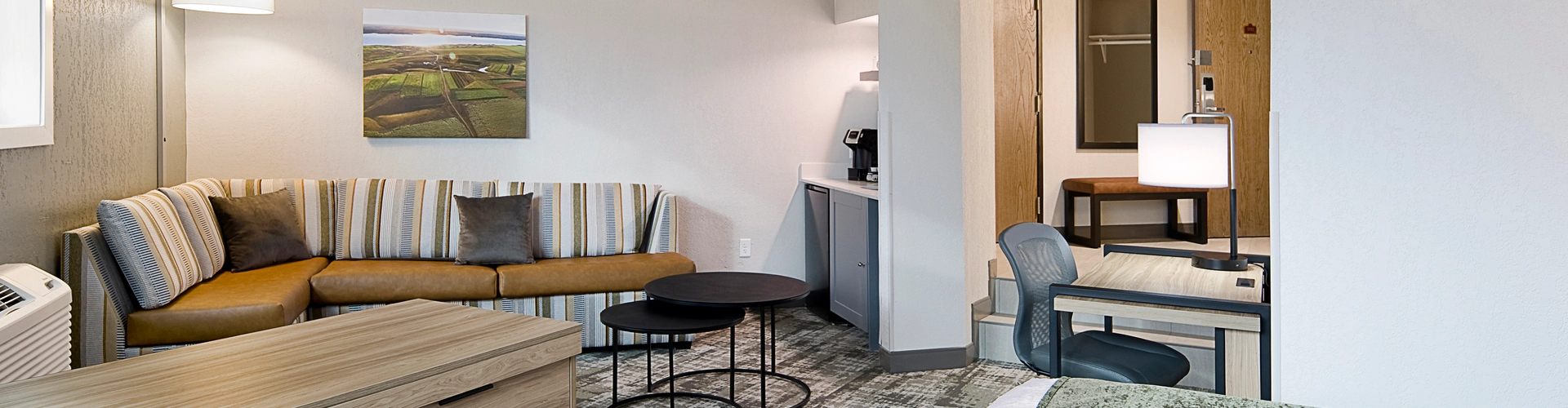 Hotel room with a modern design featuring a bed with a green throw, a wooden dresser, a sofa with striped cushions, a small table, and a workstation with a chair. The walls are decorated with a landscape photo.