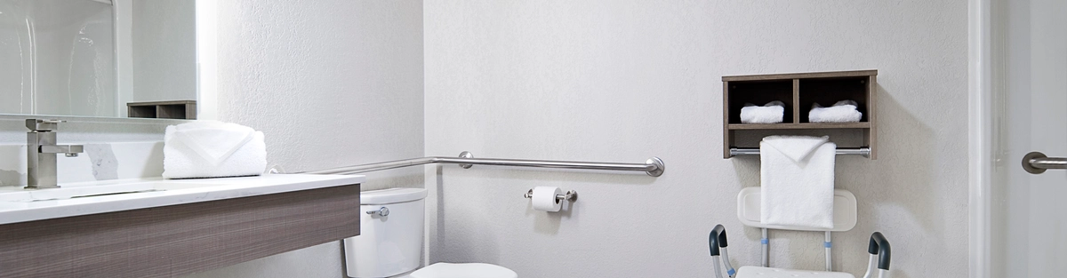 A spacious, accessible bathroom with a white toilet, folding shower chair, and safety grab bars. A modern vanity holds a rolled white towel. Neutral-toned walls and floor tiles create a minimalist, clean appearance.